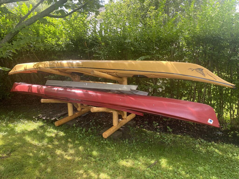 Customer's log kayak rack storing 2 kayaks and a ladder on top.  Some bushes are in the background. 