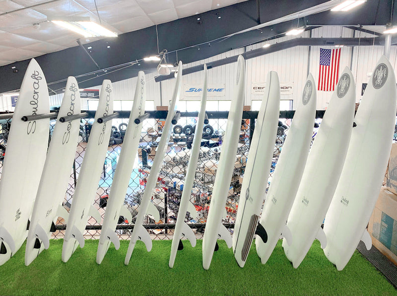 Front view of two black metal surf racks mounted to a fence overlooking a boating showroom floor.  The racks are holding several white wakesurf boards in the vertical position.  The boards are laying on fake grass turf.