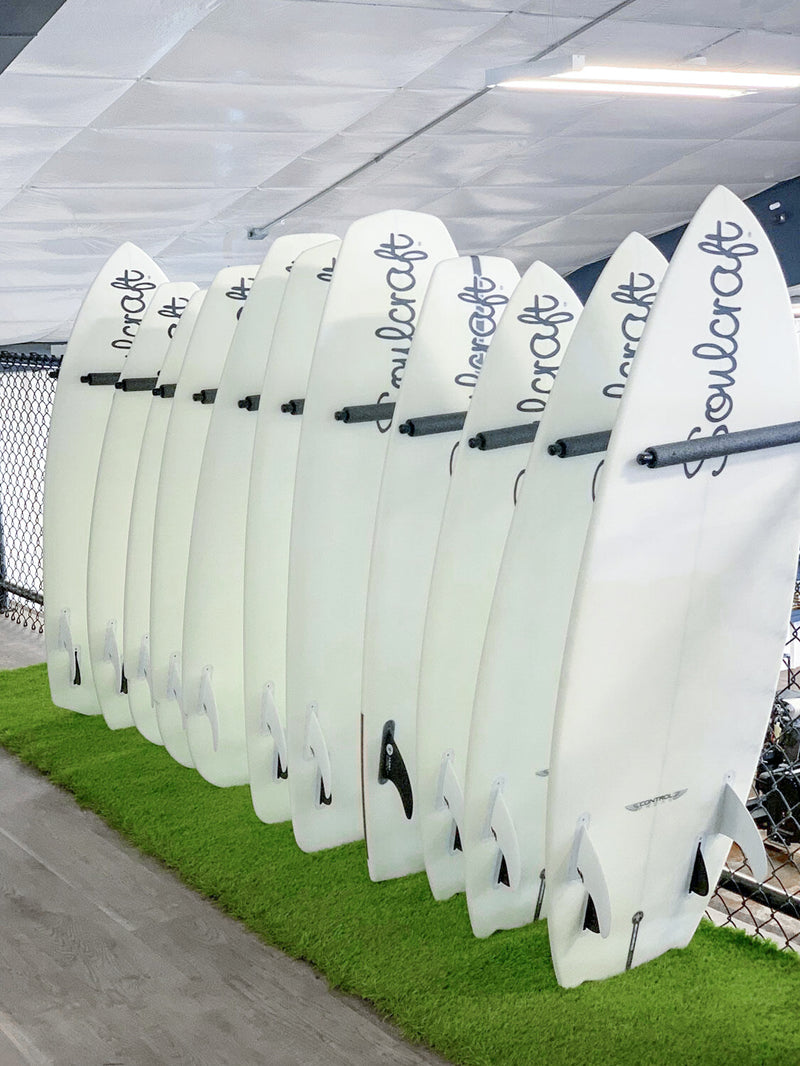 Black metal surfboard wall rack holding several Wake Surf Boards in the vertical upright position.  The boards are laying on top of faux grass.