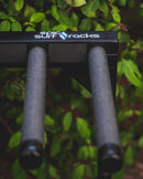 Upper front detail shot of the blackout grassy surfboard rack. The rack has two dowels covered in foam coming off the rack, and a sticker that says epic surf racks in white. There is a vibrant green bush in the background.
