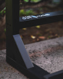 Lower left detail shot of the blackout surfboard rack showing the matte black paint. There is a cross-bar that has a white sticker that says epic surf racks. There is a bush in the background.