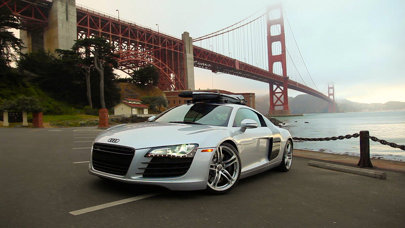 An Audi R8 with a surfboard mounted on the roof with the universal-mount suction cup roof rack. The car is silver and is parked in front of the golden gate bridge in San Francisco in the background. 