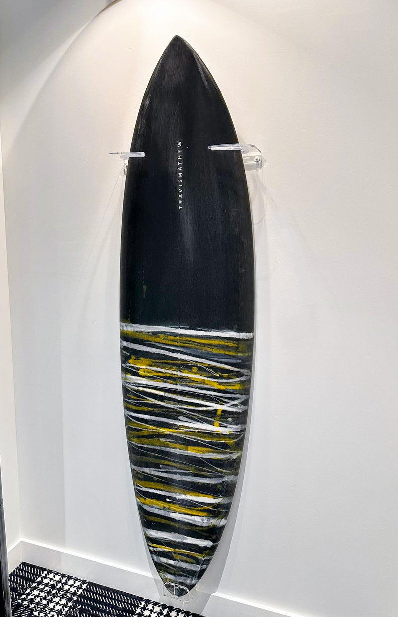 Travis Matthew surfboard being displayed in a retail setting.  The board is mounted on the wall in a vertical position with acrylic wall hangers.  The surfboard is black with white and yellow stripes on the bottom. 