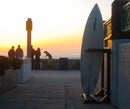 Picture of a sunset with the silhoutte of an epic surf racks vertical freestanding rack in the foreground, laying next to a wall.There is a group of people in the background next to a seawall watching the sunset.