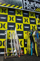 Pulled back shot of the Foamy freestanding surfboard stand holding several professional surfers boards during the Hurley Pro contest. There are several wetsuits on a rack to the right of the board rack. The hurley pro branding is all around in the background in black and yellow.