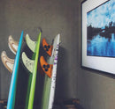 3 surfboareds shown next to each other in a vertical storage mount with the bottoms of the boards facing upright. There is a blue surfboard with wooden fins, a green surfboard with white fins, and a white surfboard with orange fins.