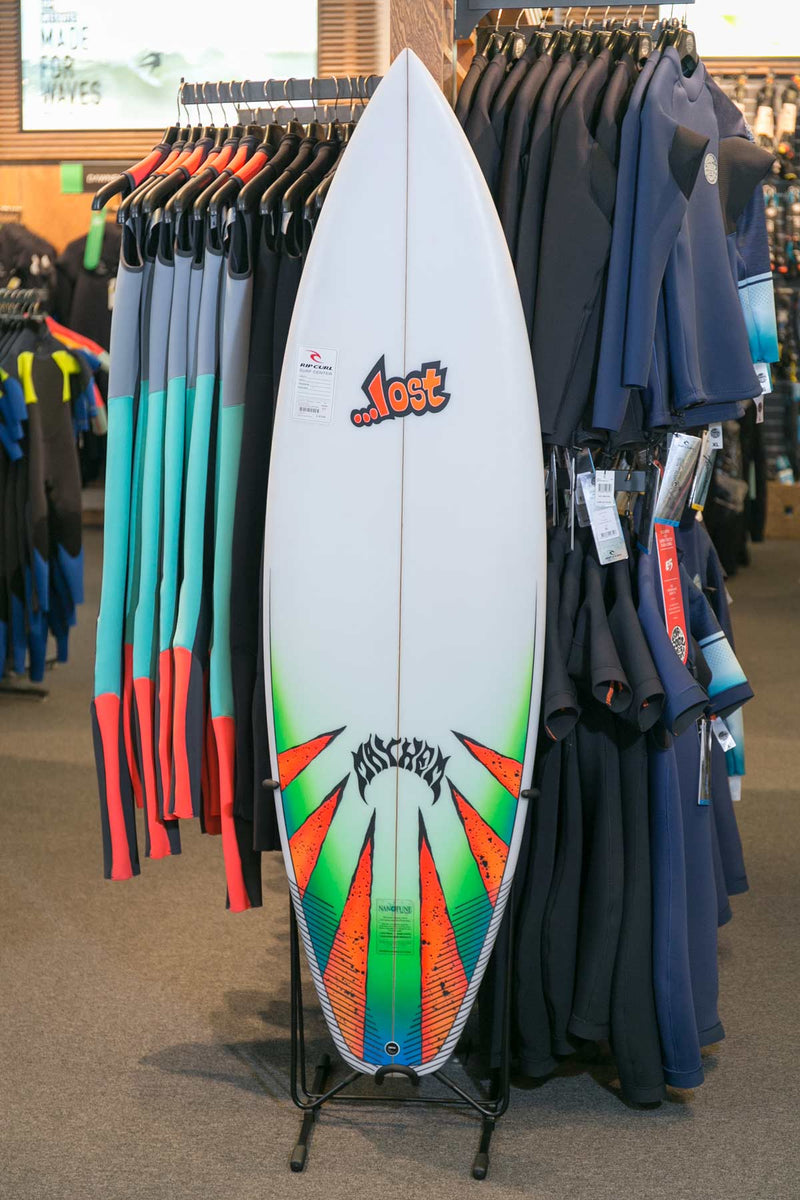 White, orange, and green shortboard surfboard being shown on display in a surf shop next to some wetsuits.  The board is standing up with the steel single freestanding surf rack.