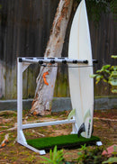 Marshmallow Grassy surf rack shown in white on the side of a yard. This vertical freestanding surfboard rack is holding just a single shortboard. The faux grass base is shown protecting the bottom of the board. There are bushes in the foreground and a tree & fence in the background.