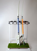 White freestanding surfboard rack made out of wood for 5 surfboards. The rack is holding two surfboards while they lay on the green grass bottom that protects the boards.