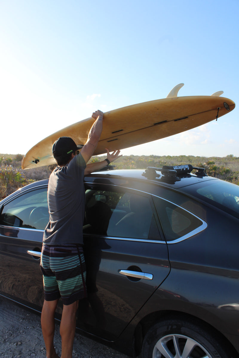 Picture of a man putting a surfboard on a suction mount roof rack that is attached to a black sedan.  The roof rack is mounted via suction cups. 