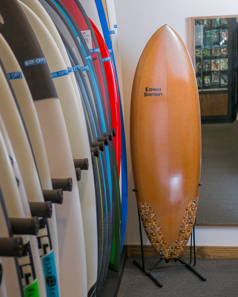 Steel Single Freestanding Surfboard rack shown in the corner of a surf shop surrounded by boards.  The Single Freestanding rack is holding a brown & gold surfboard.  
