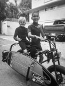 Two kids on an e-bike electric motor driven bicycle with a surfboard attached. The kids are in wetsuits are smiling and giving the shaka gesture with their hands.