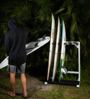 Marshmellow foam freestanding surfboard rack holding multiple surfboards in front of some palm plants, while a surfer holds a board and walks off away from it.