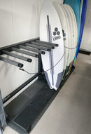 blackout foamy vertical freestanding surfboard rack in matte black shown in a customers garage. 2 racks are sitting next to one another. One rack is empty, and the other is holding about 9 surfboards. The walls are white, and the floor is concrete.