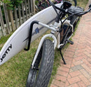Closeup view of the U-bolt & frame mount e-bike surfboard rack on a white beach-cruiser style electric bike. The bike's surf rack is holding a white lost surfboard.