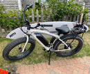Side view of the U-bolt & frame mount surfboard rack for e-bikes on a beach cruiser styled electric bike. The bike's rack is holding a white board. The bike has fat tires.