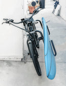 Black electric bicycle shown leaning against a white wall. The black bike has a surf bike rack, holding a blue longboard with a single fin.