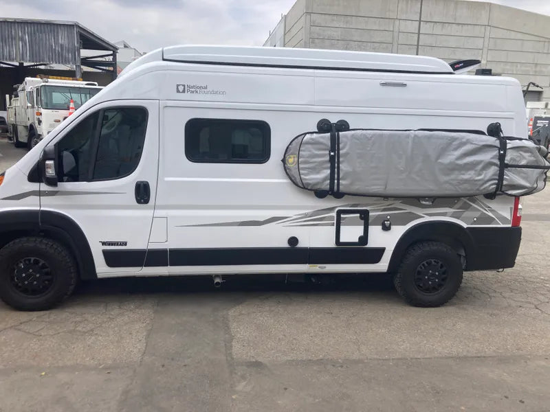 White camper van with a surfboard mounted on the side.  The surfboard is in a grey board bag.  The rack is being held to the van with suction cups. There is a grey concrete building in the background. 