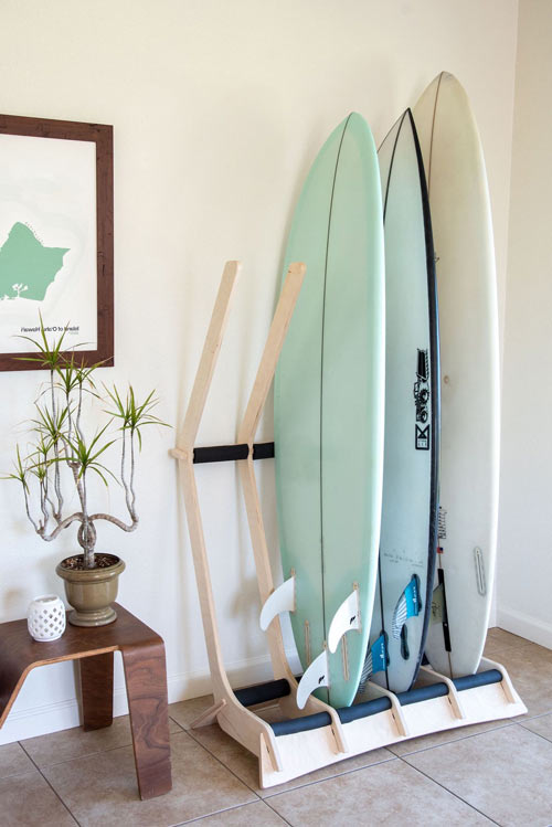 4-board freestanding floor rack shown in a living room next to a wooden table.  The surfboard rack is a light colored wood, and is holding 3 surfboards with an empty slot for an additional surfboard.  