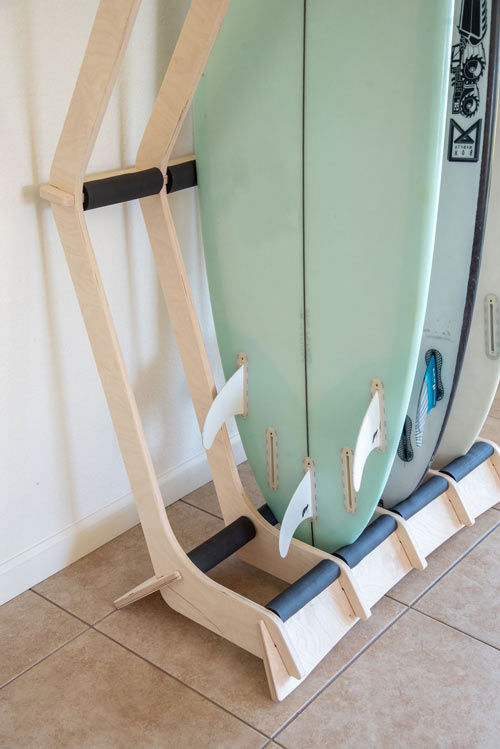 Bottom of the 4-board wooden freestanding surf rack tail and rail protection.  The rack is sitting on brown tile, and is holding several surfboards. 