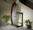 blackout grassy freestanding rack in living room stacked with surf boards.There are house plants to each side of the flat black surfboard rack. The rack is sitting on grey wooden floors, with white trim