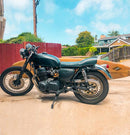 Surfboard Moped Rack | Dual-Mount -shown mounted on a motorcycle and holding a yellow longboard. The bike is parked in a driveway with houses in the background.