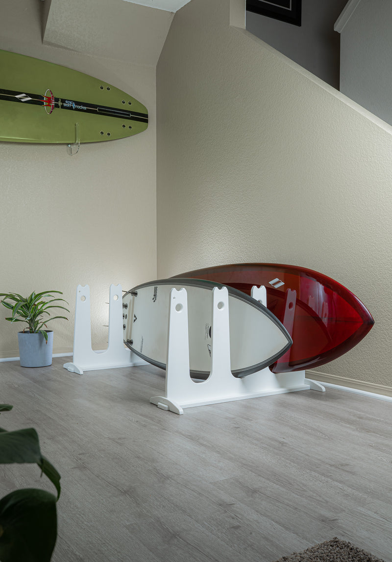 White SUP and Surfboard Dock Rack shown holding 2 surfboards next to a staircase inside a home. Another green surfboard is mounted on the wall in the background with some plants on the floor and in the foreground.  There is grey wooden floors.