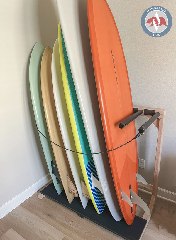 Vertical freestanding surfboard rack loaded with a variety of colorful surfboards in the corner of a room.  There is a logo at the top right of the image that says Hand-Made USA