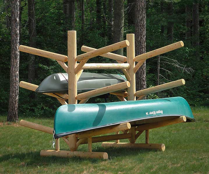 6 SUP and Kayak Log Storage Rack shown out in the woods.  The rack is sitting on grass with trees in the background.  The 6 level wooden rack is holding a Canoe on one side, and a kayak on the other. 