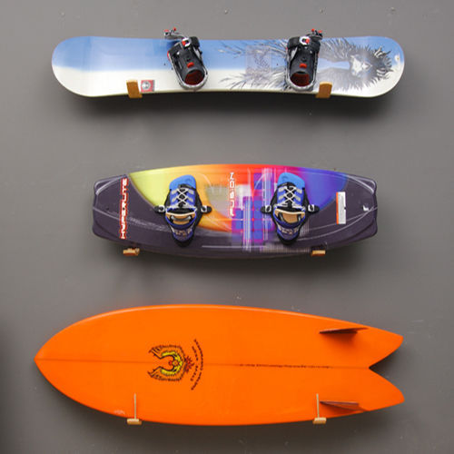 3 of the Wooden Surfboard Wall Racks mounted directly above each other.  One rack is holding a snowboard.  Below this a similar rack is holding a dark blue wakeboard, below this the same type of rack is holding an orange fish surfboard. All three of these racks are mounted to a dark grey wall. 
