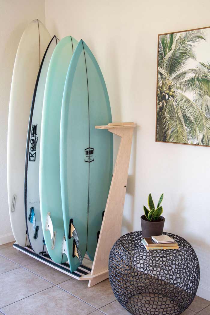 Premium 4-surfboard board freestanding floor rack in use in a nicely lit home with other furniture items.  There is a picture of palm trees next to the surf rack. 