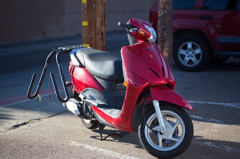 Motorcycle & Moped Surfboard Rack Single mount shown on a red moped parked in a parking lot with telephone poles in the background.