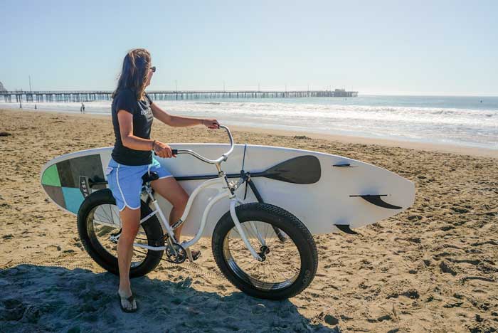 SUP Bike rack shown being used by a woman at the beach. She is checking the surf while her Stand Up Paddle Board is mounted to her white beach cruiser. There is a pier in the background.