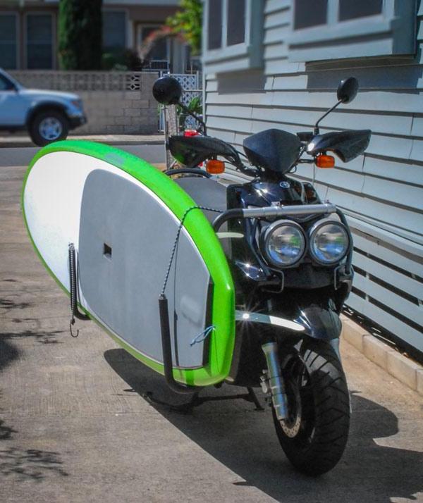 Moped shown holding a green Stand Up Paddleboard. The bike is parked in a driveway under a car port.