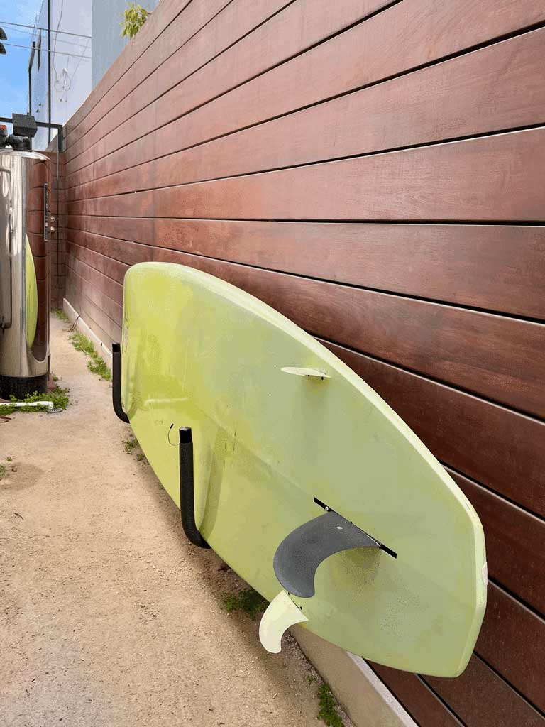 Stand UP Paddle board wall cradle rack being used in a customers backyard.  The SUP rack is attached to a wooden fence on the side of a house.  The board is yellow and appears to be quite large. 