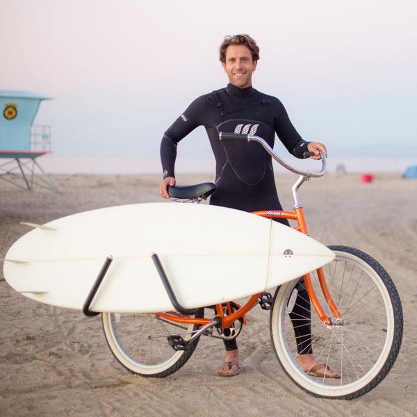 Single-Mount surfboard bike rack shown on the beach attached to an orange beach cruiser.  A man in a wetsuit is behind the bicycle and is smiling. 