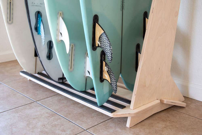 Detail of rubber tail protection being used by multiple surfboards.  Showing Rounded Pins and a Fish surfboard.   The surfboards are shown with their fins installed.  The surf rack is made of wood.  The floor it sits on is made of tile. 