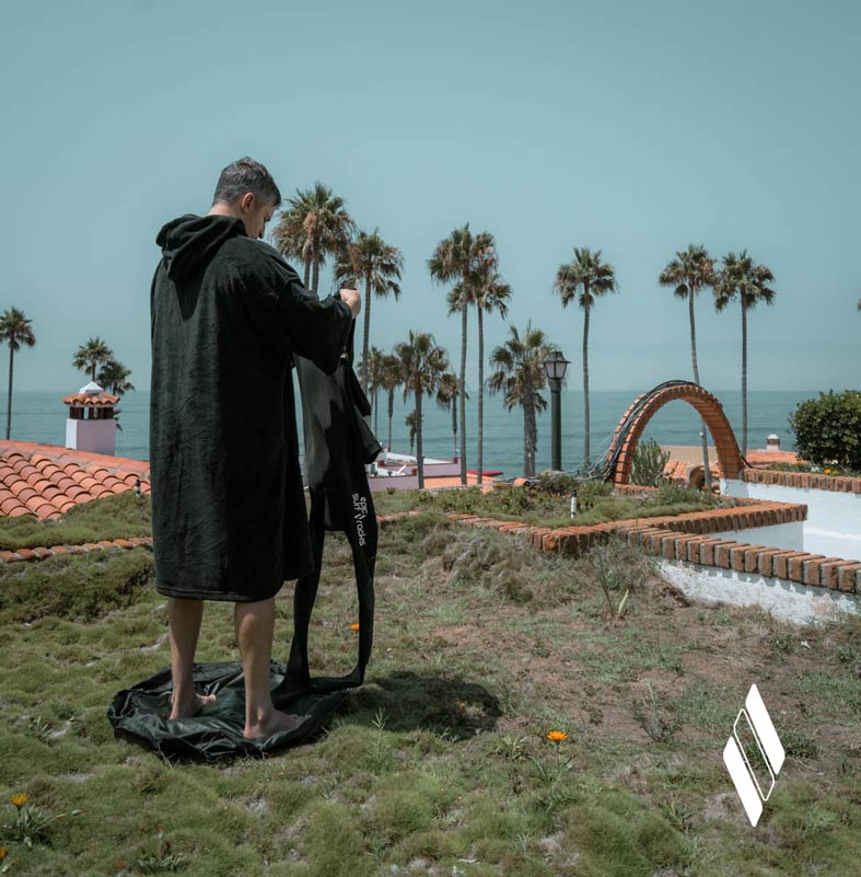 Epic Surf Racks black microfiber wetsuit changing robe and changing mat being used by the beach to change into a wetsuit on a grass patch by the ocean surrounded by palm trees.