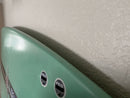 Another angle of the flush mount acrylic rack showing how close the surfboard is mounted to the wall. The surfboard is almost entirely flush with the wall. The surfboard is green, and has FCS fin plugs shown in the lower middle portion of the image.