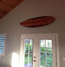 Wooden surfboard shown mounted using the acrylic flush mount surfboard holder above a doorway. There is a wooden ceiling in the room, and a deck just beyond the glass doors.