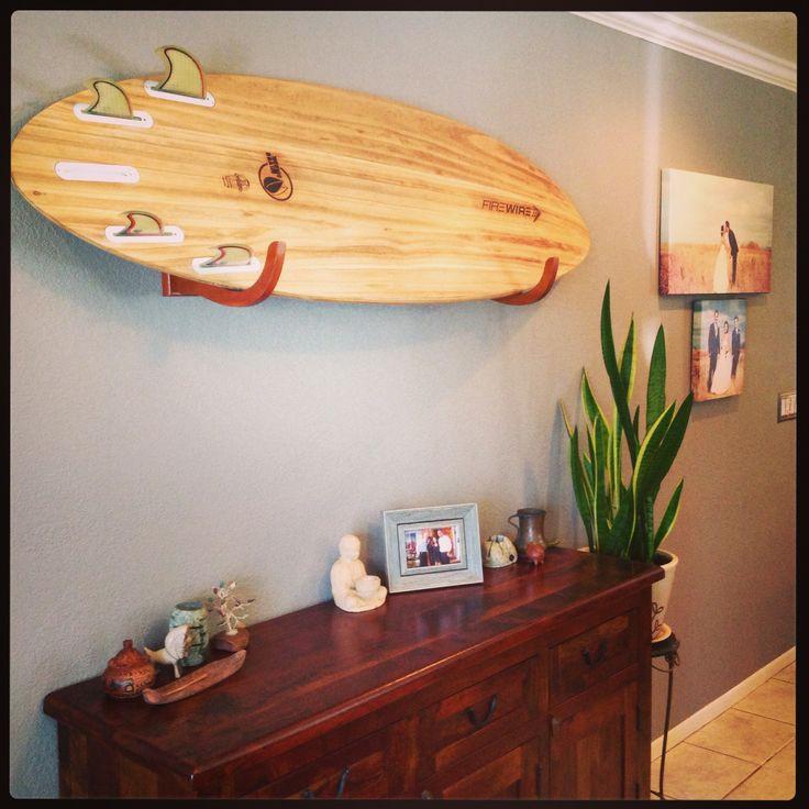 Wooden Surfboard Wall Rack shown mounted to a dark grey wall in a living room.  The board rack is holding a wooden firewire surfboard in the horizontal position. There is a dark brown wooden cabinet below where the surfboard is mounted. 