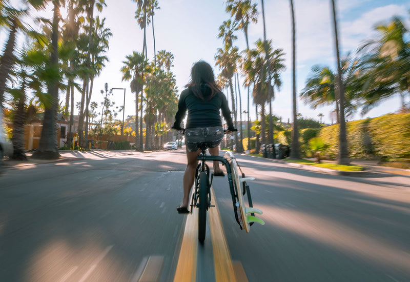 Picture of a girl riding a bike down the street, with a surfboard attached to the side of a bike using a surfboard bicycle rack.  She is passing by several palm trees, and the image is somewhat blurry showing that she might be going fast. 