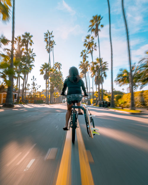 Picture of a girl riding a bike down the street, with a surfboard attached to the side of a bike using a surfboard bicycle rack.  She is passing by several palm trees, and the image is somewhat blurry showing that she might be going fast. 