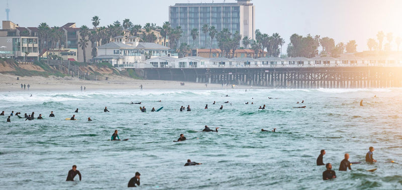 Busy Southern California surfing lineup showing many surfers battling for waves with a pier and buildings in the background.