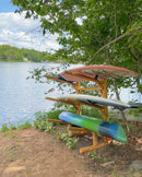 4 Kayak or SUP Log rack shown holding multiple stand up paddle boards and a kayak while the rack is sitting next to a lake with some trees in the background and off into the distance.