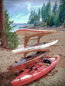 Wooden freestanding log kayak rack holding two stand up paddle boards, and two kayaks. The rack is freestanding and is on a beach next to a large lake with some trees in the background.