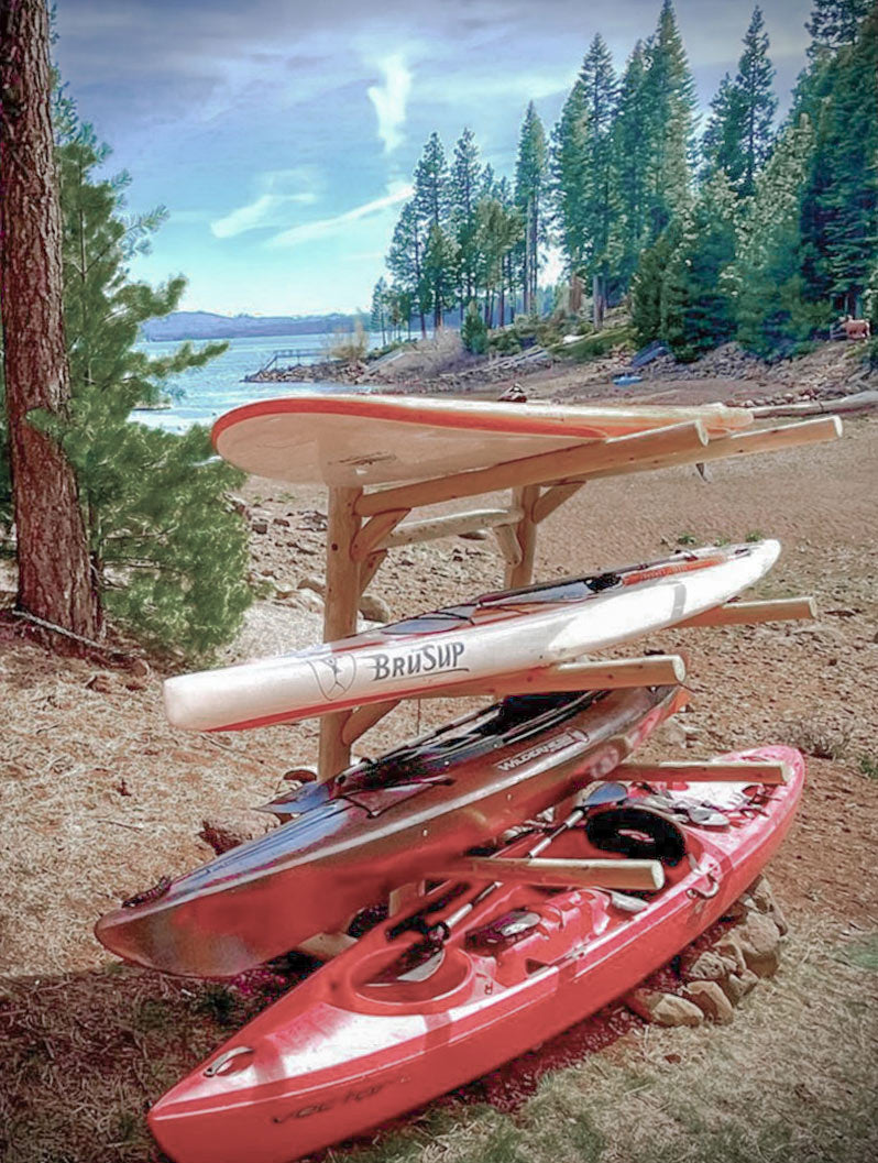 Wooden freestanding log kayak rack holding two stand up paddle boards, and two kayaks. The rack is freestanding and is on a beach next to a large lake with some trees in the background.