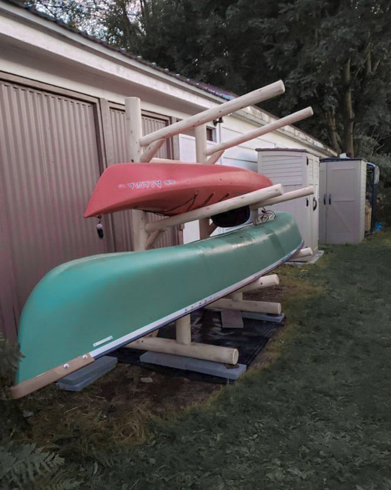4 level log kayak rack (single-sided) standing next to a garage and storage sheds.  The log kayak rack is holding both a green Canoe and a red Kayak