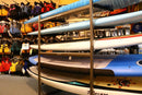 Freestanding SUP rack shown in a surf shop in front of a bunch of life vests. The SUP rack is holding several watercraft. 