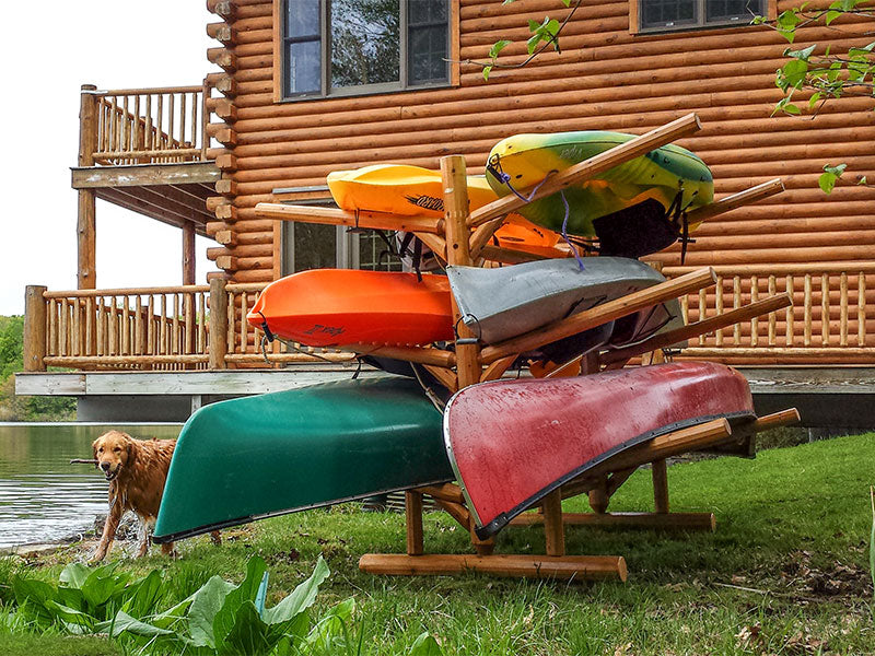 Log Kayak and Canoe rack on the side of a lake in front of a cabin.  A golden retriever dog is off to the left of the rack.  The rack is holding several kayaks and canoes